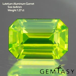 May include: A bright, rectangular-cut, green Lutetium Aluminum Garnet gemstone. The gem measures 6x4mm and weighs 1.27 ct. The gemstone is faceted, reflecting light, and is set against a grey background. The image includes the Gemtasy logo.