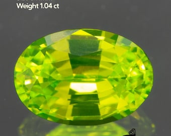 May include: A vibrant, oval-cut Lutetium Aluminum Garnet gemstone. The gem is a bright, translucent green with a faceted surface. Text indicates the size is 6x4mm and weighs 1.04 ct. The Gemtasy logo is in the bottom right corner.
