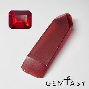 Puede incluir: Una gema rectangular facetada de color rojo vibrante con bordes afilados y acabado pulido, junto a una punta de cristal larga, translúcida y de color rojo intenso con base plana, que incluye el texto GEMTASY en la parte inferior.