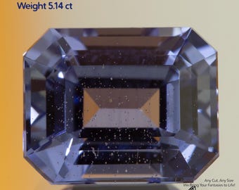 May include: A rectangular, light blue gemstone with an emerald cut. The text on the image reads "Grown Sapphire Cornflower Light, Size 10x8mm, Weight 5.14 ct." The gem is set against a gradient background.