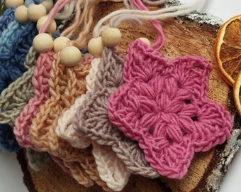 Op de afbeelding: Een set van zeven gehaakte sterdecoraties in verschillende kleuren, waaronder roze, bruin, beige en blauw. Elke ster is bevestigd aan een houten kraal en een lengte garen.