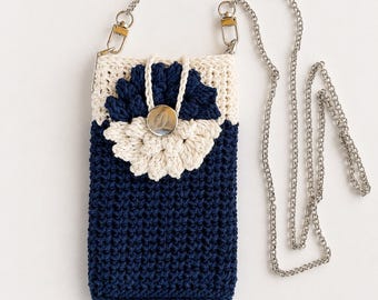 May include: A rectangular crochet crossbody bag featuring a deep navy blue body and a cream-colored top section. A layered, circular crochet flower in navy and cream sits on the front, secured by a silver button. It has a silver metal chain strap.