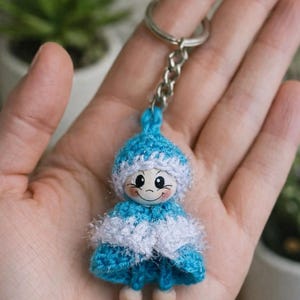 Op de afbeelding: Een handgemaakte sleutelhanger met een kleine, gehaakte pop. De pop heeft een blauw-witte outfit, een lachend gezicht en houten voetjes. De sleutelhanger is bevestigd aan een zilveren ketting en ring.