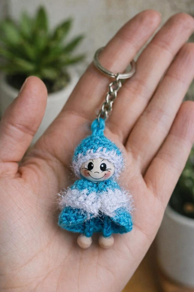 Op de afbeelding: Een handgemaakte sleutelhanger met een kleine, gehaakte pop. De pop heeft een blauw-witte outfit, een lachend gezicht en houten voetjes. De sleutelhanger is bevestigd aan een zilveren ketting en ring.