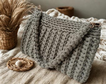 May include: A sage green crochet handbag featuring a thick, textured knit pattern with a fold-over flap design. The bag is equipped with metallic gold-toned hardware, including side clasps for a detachable strap, showcasing a dense, uniform stitch.