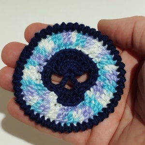 May include: A crocheted skull design with a blue skull and a white, blue, and purple background.