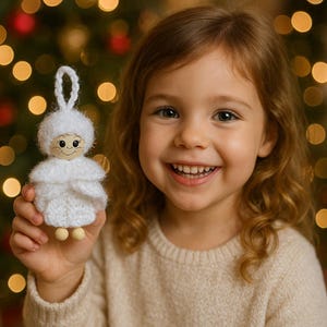 Op de afbeelding: Een lachend kind houdt een handgemaakt wit engelenornament vast. De engel heeft een houten hoofd met geschilderde kenmerken, een gebreid lichaam en kleine houten voetjes. De achtergrond toont vage kerstboomlichtjes.