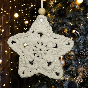 Può includere: Un ornamento a forma di stella in crochet bianco appeso a un albero di Natale con decorazioni dorate e nere. La stella ha un design delicato e traforato.