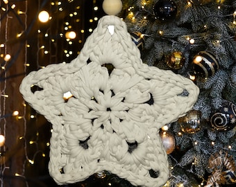 Op de afbeelding: Een witte gehaakte sterversiering die aan een kerstboom hangt met gouden en zwarte kerstballen. De ster heeft een delicaat, kantwerkontwerp.