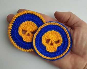 May include: Two crocheted skull-shaped coasters with a blue background and a yellow skull design. The coasters have a yellow border.