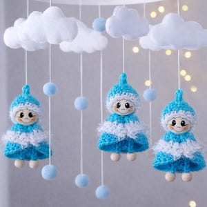 Op de afbeelding: Een handgemaakte baby mobile met drie lachende poppen in blauw en witte gehaakte jurken en hoeden, hangend onder witte vilten wolken en blauwe pompons. De mobiel hangt aan een wit cirkelvormig frame.
