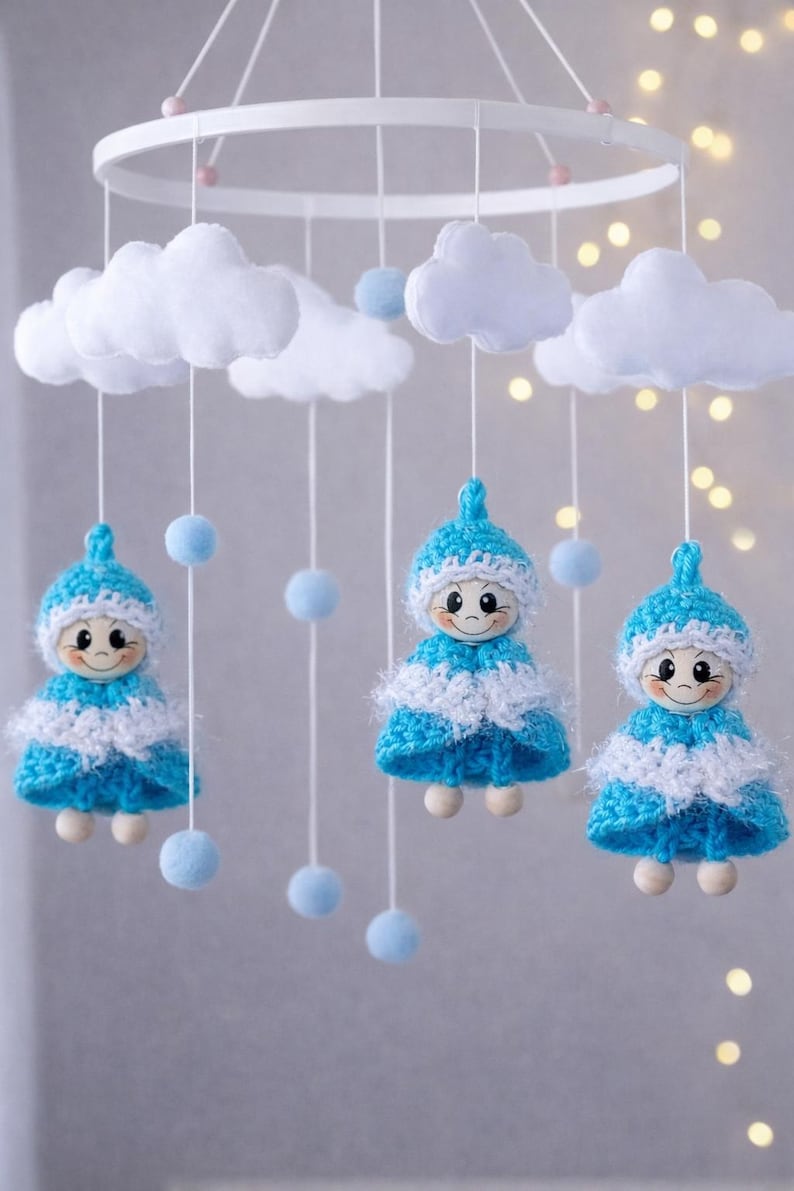 Op de afbeelding: Een handgemaakte baby mobile met drie lachende poppen in blauw en witte gehaakte jurken en hoeden, hangend onder witte vilten wolken en blauwe pompons. De mobiel hangt aan een wit cirkelvormig frame.