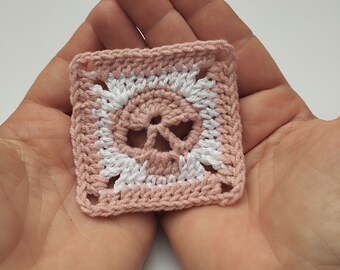 May include: A pink and white crocheted square with a skull design in the center.