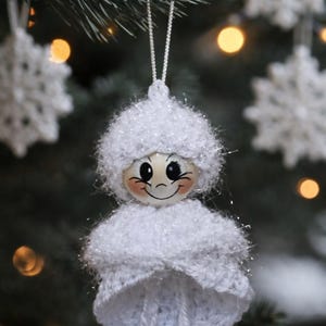 Op de afbeelding: Een handgemaakte kerstboomversiering in de vorm van een lachende engel. De engel is gemaakt van wit garen en heeft een geschilderd houten gezicht met roze wangen. De versiering hangt aan een zilveren draad, met een vage achtergrond van een kerstboom.