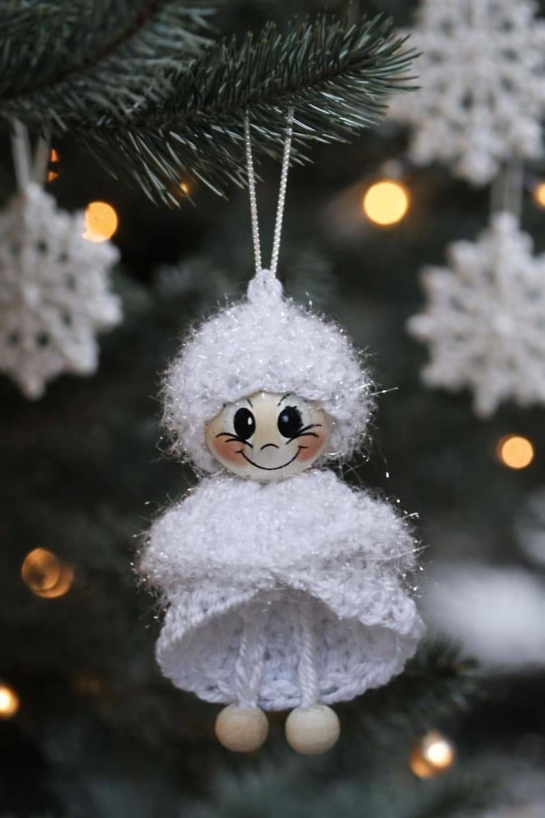 Op de afbeelding: Een handgemaakte kerstboomversiering in de vorm van een lachende engel. De engel is gemaakt van wit garen en heeft een geschilderd houten gezicht met roze wangen. De versiering hangt aan een zilveren draad, met een vage achtergrond van een kerstboom.