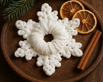 Op de afbeelding: Een witte, pluizige, gehaakte sneeuwvlokversiering ligt op een houten bord met gedroogde sinaasappelschijfjes en kaneelstokjes. Een takje groenblijvend voegt een vleugje groen toe. De sneeuwvlok heeft een cirkelvormig midden en zes punten.