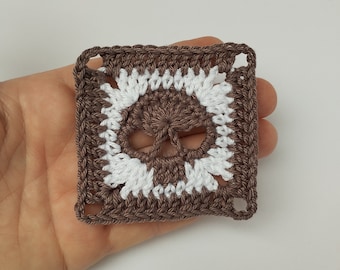 May include: A brown crocheted square with a white crocheted skull design in the center.