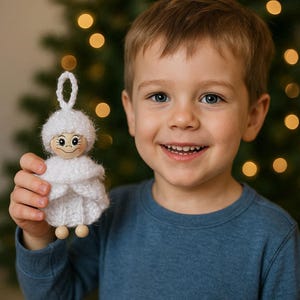 Op de afbeelding: Een handgemaakte witte gebreide engel kerstversiering met een houten hoofd en kralenvoeten. De engel heeft een lus om op te hangen en wordt vastgehouden door een lachend kind. De achtergrond heeft een vage kerstboom met warme lichten.