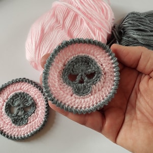 May include: Two crocheted skull appliques with a pink and gray color scheme. The skull is gray with a pink background and a gray border.