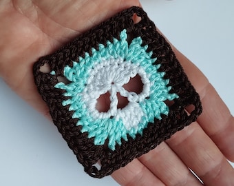 May include: A crocheted square with a skull design in white and turquoise yarn. The skull is surrounded by a brown border.