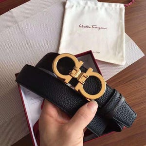 Puede incluir: Cinturón de cuero negro con hebilla dorada. La hebilla presenta un logotipo de doble G. El cinturón se muestra en una caja roja con una bolsa blanca con cordón en el fondo. La bolsa tiene el nombre de la marca impreso.