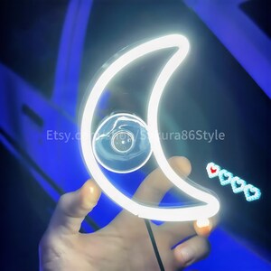 Puede incluir: Un letrero de luna creciente de neón blanco con una ventosa en la parte posterior. El letrero está siendo sostenido por una mano contra un fondo azul.