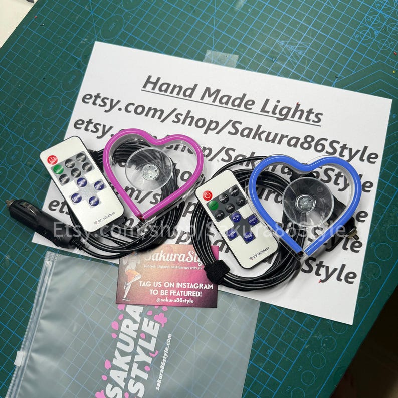 May include: Two heart-shaped LED lights with remote controls. The lights are pink and blue and have suction cups for attaching to a car window. The lights are connected to a car charger. The image also includes a business card with the text "Sakura Style" and instructions to tag the business on Instagram.