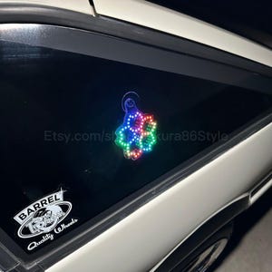 Puede incluir: Una luz LED de colores arcoíris con forma de flor está adherida a la ventana trasera de un coche blanco. La luz está adherida con una ventosa. El texto "Etsy.com/shop/sakura86Style" es visible en la ventana.