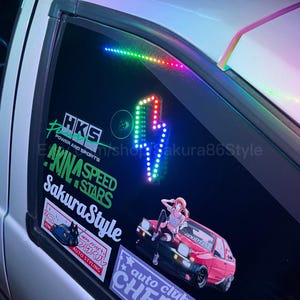 May include: A white car with a variety of colorful stickers on the back window. The stickers include the text "HKS", "AKINA SPEED STARS", "SakuraStyle", "GOOD LINE", and "auto club CHE".