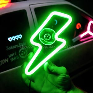 May include: A green neon lightning bolt sign with a white outline. The sign is held in a hand and is lit up against a dark background.