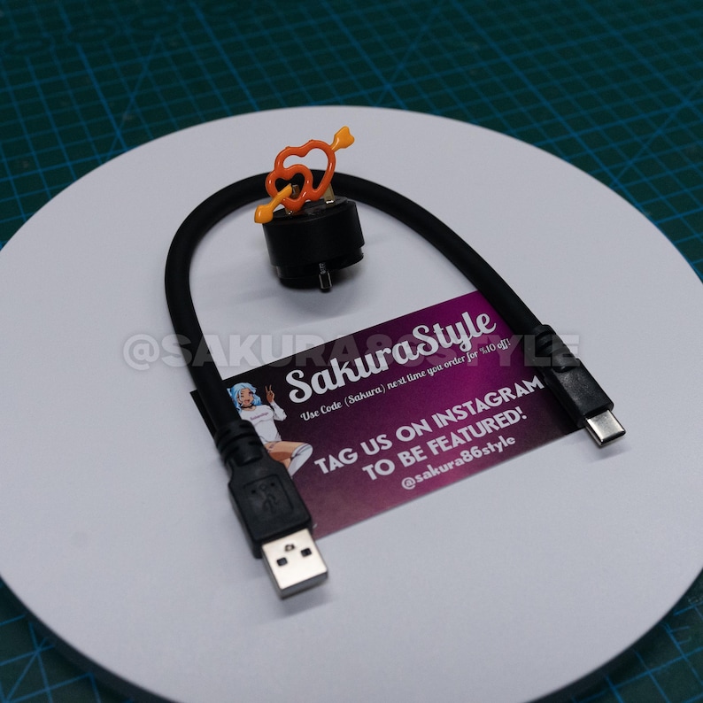 May include: A black USB charging cable with a USB-A connector and a USB-C connector. The cable has a black base with an orange heart-shaped charm. A business card with the text "SakuraStyle" is also present.