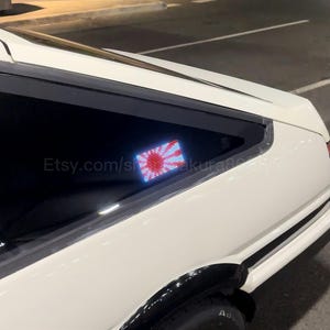 Pode incluir: Um carro branco com acabamentos pretos, exibindo um autocolante de janela com o design da bandeira japonesa. O autocolante mostra um sol vermelho e raios vermelhos e brancos. O nome da loja Etsy é visível na janela.