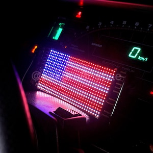 Könnte beinhalten: Ein Auto-Armaturenbrett mit einer elektronischen Anzeige, die die amerikanische Flagge in roten, weißen und blauen LED-Lichtern zeigt. Der Tachometer zeigt 0 km/h an. Weitere Anzeigen und Indikatoren sind sichtbar.
