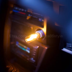 May include: A close-up of a lit, amber-colored light bulb in a car's dashboard. The bulb is in a white socket, emitting a warm glow. The dashboard is dark with visible gauges and the text "Etsy.com/sakura86style".