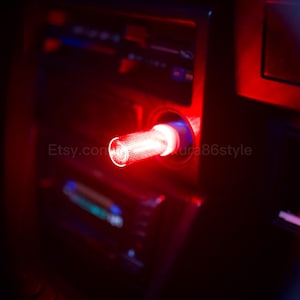 Op de afbeelding: Een rood LED-licht geïnstalleerd in de sigarettenaanstekerpoort van een auto. Het licht is cilindrisch met een gestructureerd oppervlak en straalt een heldere rode gloed uit. De tekst "Etsy.com/ura86style" is zichtbaar.