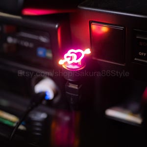 May include: A glowing pink USB car charger featuring a heart pierced by an arrow. The charger is plugged into a car's console, with the Etsy shop name "Sakura86Style" visible. The heart and arrow design emits a soft, warm light.