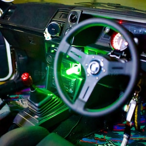 Op de afbeelding: Het interieur van een auto met een zwart stuurwiel, een zwarte versnellingspook en groene LED-verlichting. De auto heeft een zwart dashboard en een zwarte vloermat.