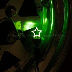 May include: A green star-shaped light, possibly a tyre valve cap, glows brightly against a shiny, metallic wheel. The star is mounted on a black base. The Etsy shop name is visible.