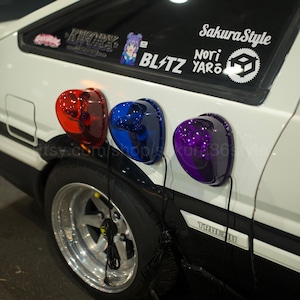 May include: A white car with black accents features three colored lights: red, blue, and purple. The car has a black tire and a silver rim. Stickers on the window read "SakuraStyle", "BLITZ", and "Nori Yaro".