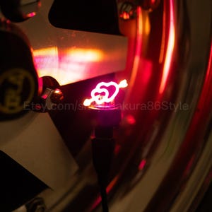 May include: A red, illuminated heart and arrow design on a tyre valve stem cap. The image is set against a blurred background of a car wheel and rim, with the Etsy shop name visible.