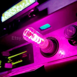 May include: A pink LED light bulb with a spiral filament is plugged into a car's cigarette lighter. The light is on and illuminating the surrounding area.