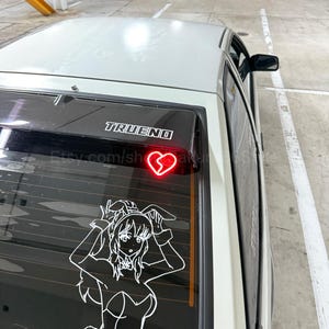 Pode incluir: Um carro branco com um spoiler traseiro de janela preto. O spoiler tem um adesivo de coração partido vermelho e a palavra "TRUENO" em letras pretas. A janela traseira também tem um adesivo de contorno branco de uma mulher em um traje de coelho.