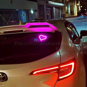 Puede incluir: Un coche blanco con una pegatina de corazón de neón rosa en la ventana trasera. El coche tiene un alerón y luces traseras rojas. El texto "Etsy.com/shop/Sakura8Style" es visible en la ventana trasera.