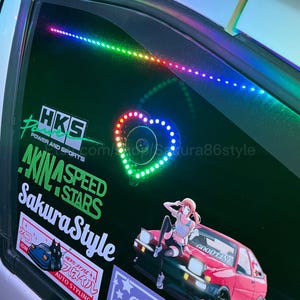 Pode incluir: Um vidro de carro preto com uma faixa de luz LED arco-íris e uma luz LED em forma de coração. O vidro também tem vários adesivos, incluindo um que diz "SakuraStyle" e outro que diz "AKINA SPEED STARS".