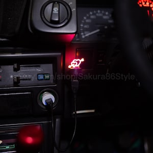May include: A red, heart-shaped USB car charger with a neon glow. The charger is plugged into a car's dashboard, with a black USB cable. The car's interior is dark, with other illuminated controls visible.