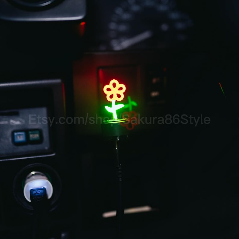 May include: A flower-shaped LED light, glowing red and green, inside a car. The text "Etsy.com/she_sakura86Style" is visible. The image is dark, with the light as the focal point.