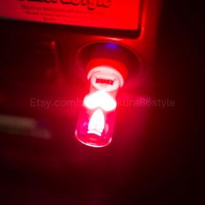 May include: A cylindrical car charger adapter emitting a bright red glow, featuring a translucent body with a white heart-shaped icon and a flame-shaped icon illuminated from within, plugged into a dark vehicle dashboard console.