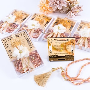 Op de afbeelding: Een set van zes gouden geschenkdozen met witte deksels, elk met een roze gebedskralenketting en een decoratieve bloem. De dozen zijn in een rij op een wit oppervlak gerangschikt. De dozen hebben een gouden inscriptie op de voorkant. De inscriptie luidt "SORRY FOR YOUR LOSS".