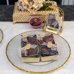 Könnte beinhalten: Vier kleine Geschenkboxen gefüllt mit Datteln und mit weißer Spitze gebunden. Die Boxen befinden sich auf einem durchsichtigen Glasteller mit goldenem Rand. Die Boxen sind mit bunten Designs und dem Text "Eid Mubarak" verziert.