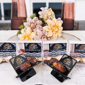 May include: Four small white boxes with clear lids contain dates and a decorative ribbon. The boxes have a black label with Arabic calligraphy and the text "In the name of Allah". Two black acrylic stands hold dates.